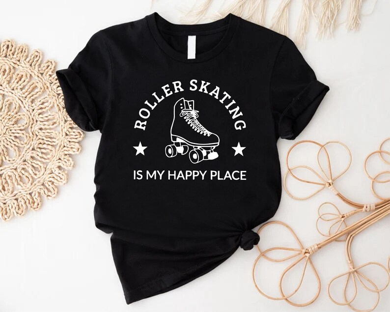 May include: Black t-shirt with a white graphic that reads "ROLLER SKATING IS MY HAPPY PLACE." The design features a roller skate and two stars. The shirt is made of a soft material and has short sleeves.
