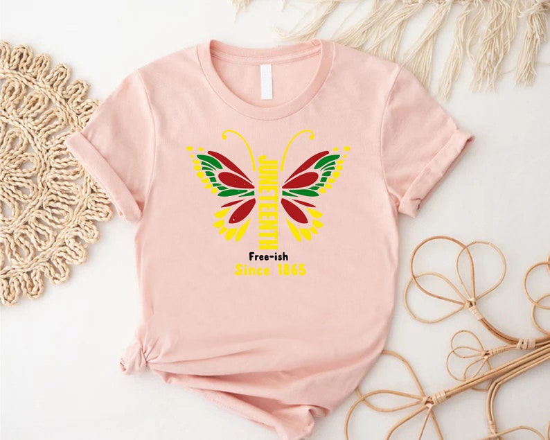May include: Pink t-shirt with a yellow butterfly graphic. The butterfly has red, green, and black wings. The text "Juneteenth" is written vertically down the butterfly's body. The text "Free-ish Since 1865" is written below the butterfly.