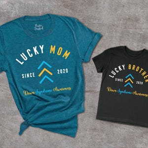 Op de afbeelding: Twee T-shirts, een tealblauw en een zwart, met de tekst "Lucky Mom Since 2020" en "Lucky Brother Since 2020" erop. Beide shirts hebben een geel en blauw naar boven wijzend pijlontwerp en de tekst "Down Syndrome Awareness".
