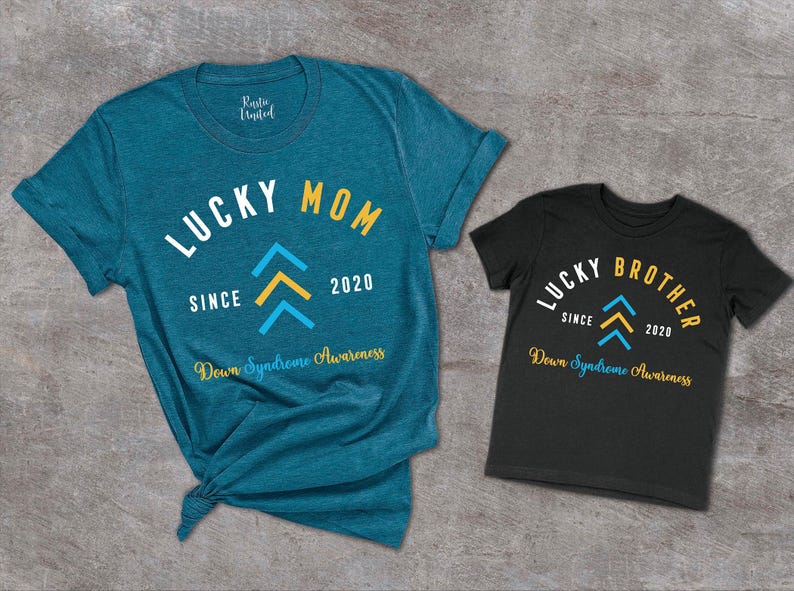 Op de afbeelding: Twee T-shirts, een tealblauw en een zwart, met de tekst "Lucky Mom Since 2020" en "Lucky Brother Since 2020" erop. Beide shirts hebben een geel en blauw naar boven wijzend pijlontwerp en de tekst "Down Syndrome Awareness".