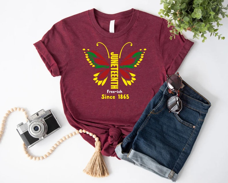 May include: A maroon t-shirt with a butterfly design featuring the words "Juneteenth Free-ish Since 1865".