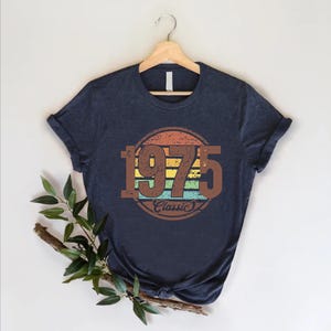 May include: Navy blue t-shirt with a retro design featuring the year "1975" in brown, set within a circular design with orange, yellow, green, and blue stripes. The word "Classic" is below the year.