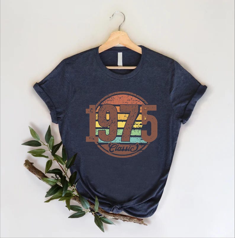 May include: Navy blue t-shirt with a retro design featuring the year "1975" in brown, set within a circular design with orange, yellow, green, and blue stripes. The word "Classic" is below the year.