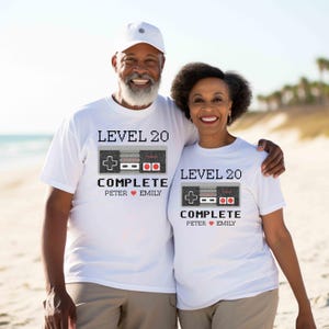 May include: Two white t-shirts with a pixelated video game controller graphic. The text on the shirts reads "Level 20 Complete" with the names "Peter" and "Emily" below.