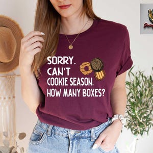 May include: A maroon t-shirt with the text "SORRY. CAN'T COOKIE SEASON. HOW MANY BOXES?" and illustrations of cookies. The shirt is paired with blue jeans. A straw hat hangs in the background.