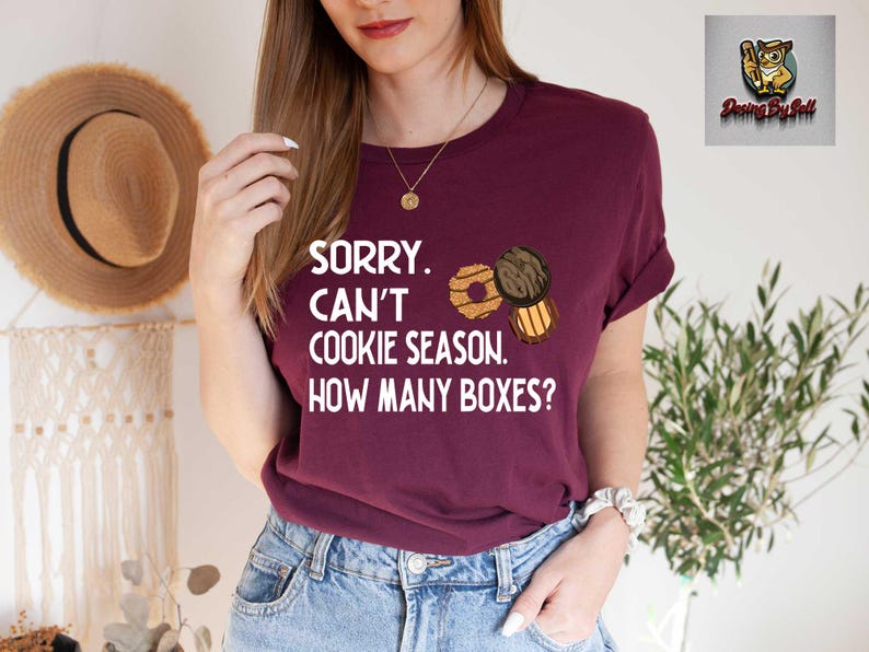 May include: A maroon t-shirt with the text "SORRY. CAN'T COOKIE SEASON. HOW MANY BOXES?" and illustrations of cookies. The shirt is paired with blue jeans. A straw hat hangs in the background.