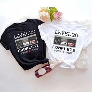 May include: Two black and white t-shirts with a pixelated video game controller graphic and the text "Level 20 Complete". The text also includes the names "Peter" and "Emily" with a heart symbol between them.