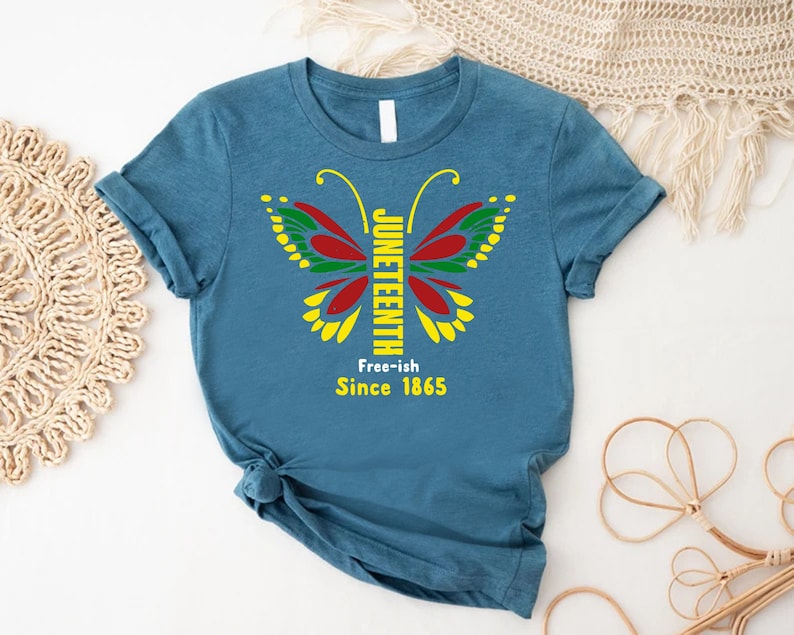 May include: A teal blue t-shirt with a butterfly graphic. The butterfly is made up of red, yellow, green, and black colors. The text "Juneteenth" is written vertically down the center of the butterfly. The text "Free-ish Since 1865" is written below the butterfly.