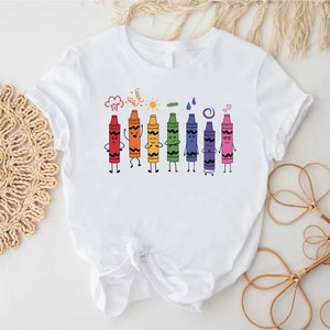 May include: White t-shirt with a colorful illustration of crayons with faces and arms. The crayons are lined up in a row and are holding hands. The text above the crayons says "Crayons are my happy place".