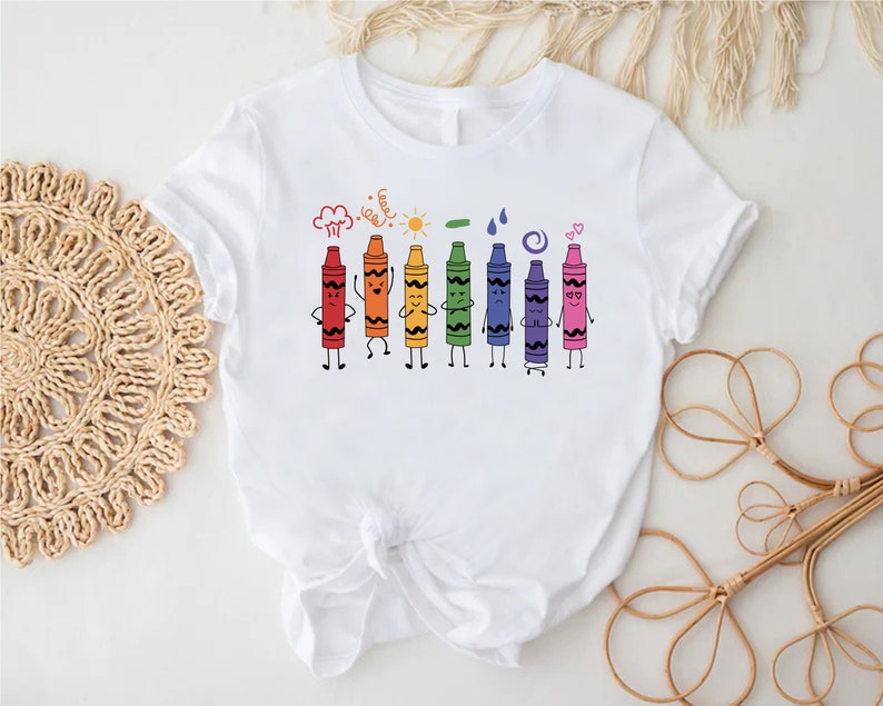May include: White t-shirt with a colorful illustration of crayons with faces and arms. The crayons are lined up in a row and are holding hands. The text above the crayons says "Crayons are my happy place".