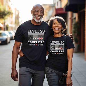 May include: Two black t-shirts with a pixelated video game controller graphic. The text on the shirts reads "Level 20 Complete" with names "Peter" and "Emily" below.