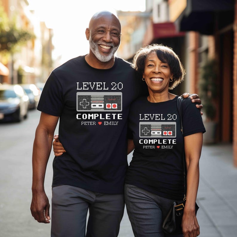 May include: Two black t-shirts with a pixelated video game controller graphic. The text on the shirts reads "Level 20 Complete" with names "Peter" and "Emily" below.
