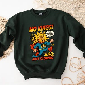 May include: Dark green sweatshirt with a cartoon graphic of a man in a superhero costume, with the text "NO KINGS! JUST CLOWNS." The man is depicted with blonde hair and a red cape. A speech bubble says "Oops... democracy wins again!"