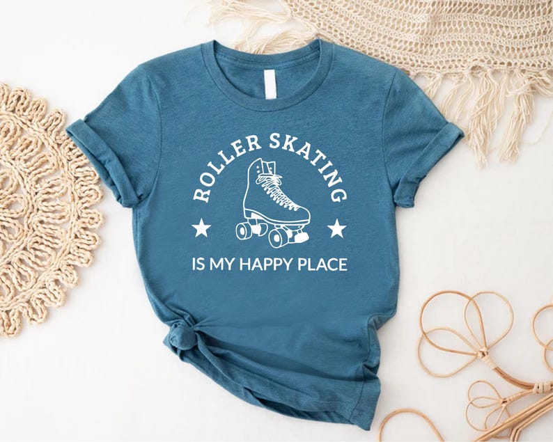 May include: Teal blue t-shirt with the text "ROLLER SKATING IS MY HAPPY PLACE" in a white, circular design. The design includes a roller skate and two stars. The shirt is made of a soft material.