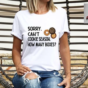 May include: White t-shirt with the text "SORRY. CAN'T COOKIE SEASON. HOW MANY BOXES?" and illustrations of cookies. The shirt is worn by a person in blue jeans, seated in a woven chair.