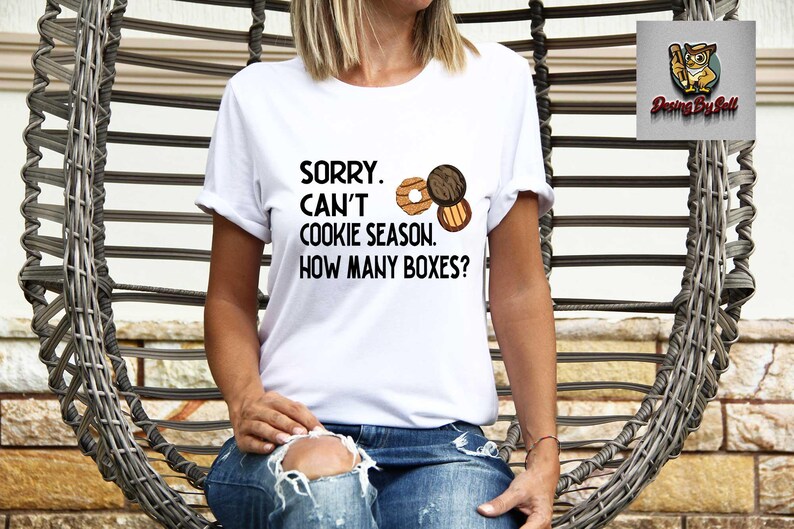 May include: White t-shirt with the text "SORRY. CAN'T COOKIE SEASON. HOW MANY BOXES?" and illustrations of cookies. The shirt is worn by a person in blue jeans, seated in a woven chair.