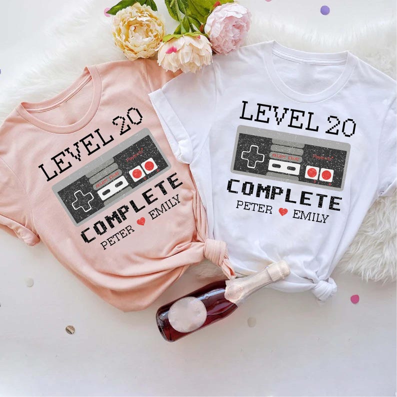May include: Two pink and white t-shirts with a pixelated video game controller graphic and the text "LEVEL 20 COMPLETE" with names "Peter" and "Emily".