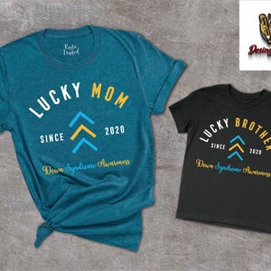 Op de afbeelding: Twee T-shirts, een tealblauw en een zwart, met de tekst "Lucky Mom" en "Lucky Brother" in wit. Beide shirts hebben een geel en blauw naar boven wijzend pijlontwerp en de tekst "Since 2020" en "Down Syndrome Awareness".