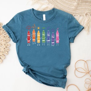 May include: A teal blue t-shirt with a graphic of a line of colorful crayons with faces. The crayons are arranged in a rainbow order, with red, orange, yellow, green, blue, purple, and pink. The crayons are smiling and have arms and legs.