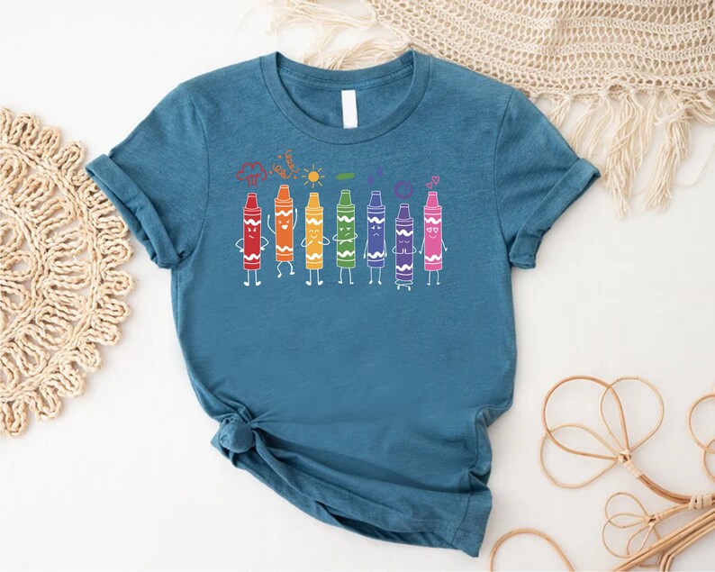 May include: A teal blue t-shirt with a graphic of a line of colorful crayons with faces. The crayons are arranged in a rainbow order, with red, orange, yellow, green, blue, purple, and pink. The crayons are smiling and have arms and legs.