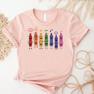 May include: A pink t-shirt with a graphic of seven colorful crayons with faces. The crayons are lined up in a row and are drawn in a cartoon style. The crayons are red, orange, yellow, green, blue, purple, and pink. The crayons are all smiling and have different expressions. The t-shirt is a great gift for teachers, students, or anyone who loves to color.