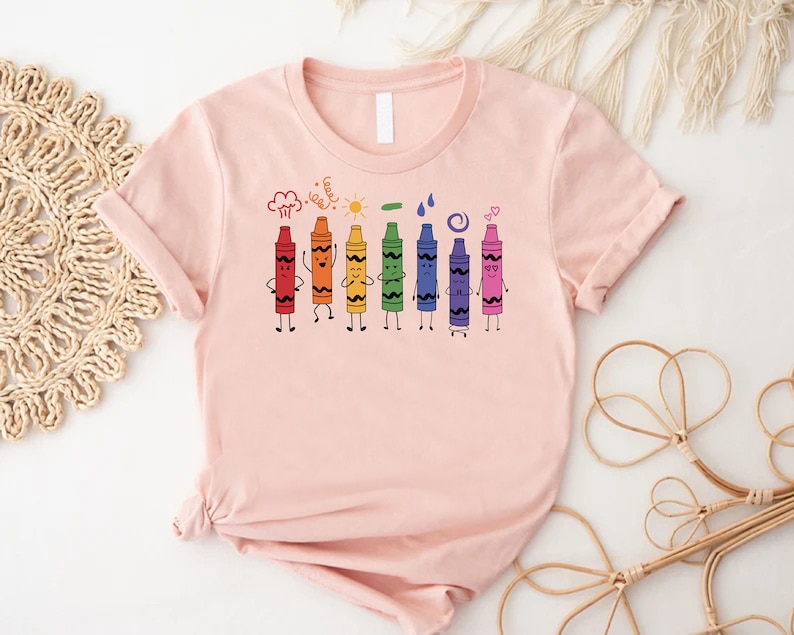 May include: A pink t-shirt with a graphic of seven colorful crayons with faces. The crayons are lined up in a row and are drawn in a cartoon style. The crayons are red, orange, yellow, green, blue, purple, and pink. The crayons are all smiling and have different expressions. The t-shirt is a great gift for teachers, students, or anyone who loves to color.