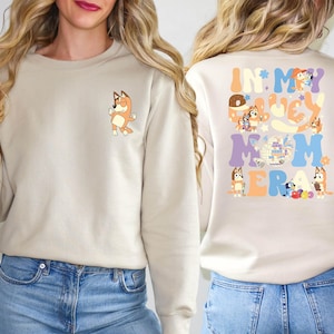 May include: Beige sweatshirt with a small cartoon character on the front and the text "IN MY LUCKY MOM ERA" with cartoon characters on the back. The design features orange, blue, purple, and yellow colors.
