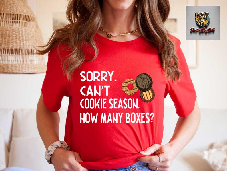 May include: A red t-shirt with the text "SORRY. CAN'T COOKIE SEASON. HOW MANY BOXES?" and illustrations of cookies. The shirt is short-sleeved and has a crew neck.