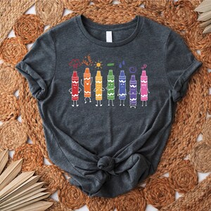 May include: A dark gray t-shirt with a design of colorful crayons with faces and arms. The crayons are lined up in a row and are holding hands. The design also includes a cloud, sun, raindrop, and hearts.