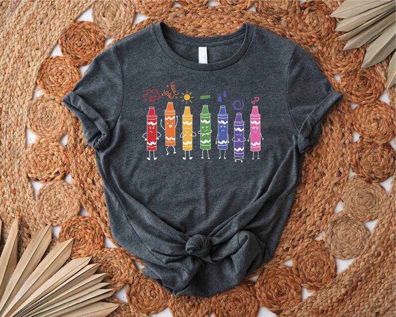 May include: A dark gray t-shirt with a design of colorful crayons with faces and arms. The crayons are lined up in a row and are holding hands. The design also includes a cloud, sun, raindrop, and hearts.