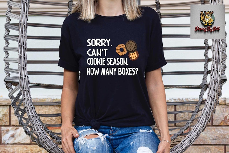 May include: Black t-shirt with the text "SORRY. CAN'T COOKIE SEASON. HOW MANY BOXES?" and illustrations of cookies. The shirt is paired with blue jeans. The design is perfect for cookie lovers.