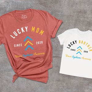 Op de afbeelding: Twee T-shirts, een rood en een wit, met de tekst "Lucky Mom" en "Lucky Brother" erop gedrukt. Beide shirts hebben een blauw en geel pijlontwerp en de tekst "Down Syndrome Awareness" gedrukt onder het ontwerp.