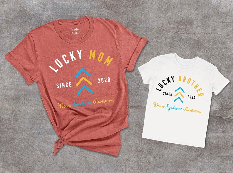 Op de afbeelding: Twee T-shirts, een rood en een wit, met de tekst "Lucky Mom" en "Lucky Brother" erop gedrukt. Beide shirts hebben een blauw en geel pijlontwerp en de tekst "Down Syndrome Awareness" gedrukt onder het ontwerp.