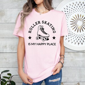 May include: A light pink t-shirt with the text "ROLLER SKATING IS MY HAPPY PLACE" in black. The design includes a roller skate illustration and star accents. The shirt is made of a soft material.