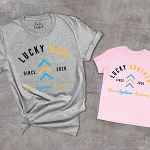 Op de afbeelding: Twee T-shirts, een grijs en een roze, met de tekst "Lucky Mom" en "Lucky Brother" erop. Beide shirts hebben een blauw en geel pijlontwerp en de tekst "Since 2020" en "Down Syndrome Awareness".