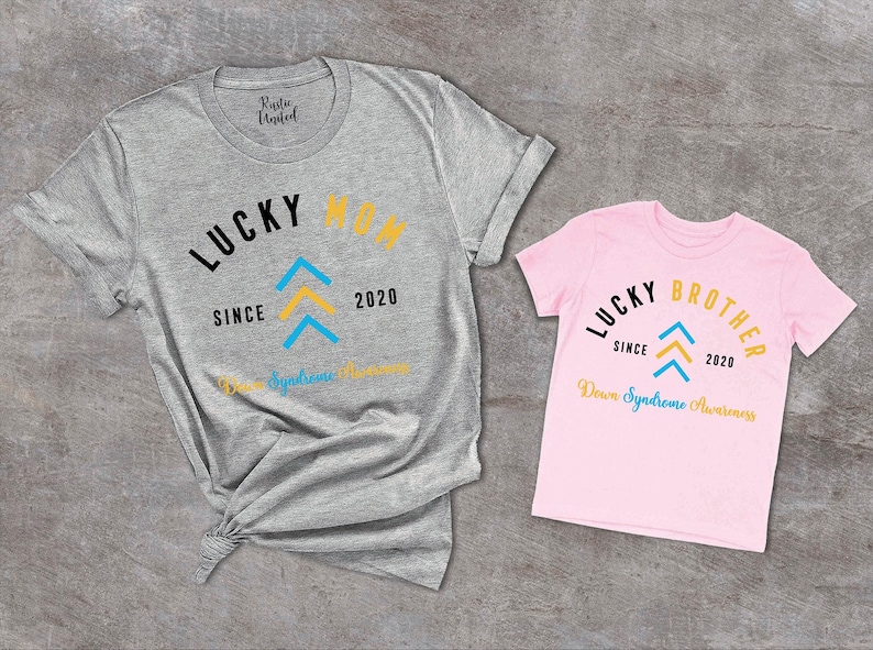 Op de afbeelding: Twee T-shirts, een grijs en een roze, met de tekst "Lucky Mom" en "Lucky Brother" erop. Beide shirts hebben een blauw en geel pijlontwerp en de tekst "Since 2020" en "Down Syndrome Awareness".