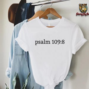 May include: A white t-shirt with the text "psalm 109:8" in black. The shirt is hanging on a wooden hanger with a light blue denim shirt and a brown hat in the background. The shirt is part of a clothing collection.