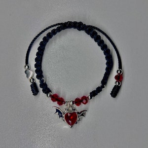 May include: A dark blue macrame bracelet with a silver heart-shaped charm featuring red accents and bat wings. The bracelet also has red beads and silver beads. The bracelet is adjustable.