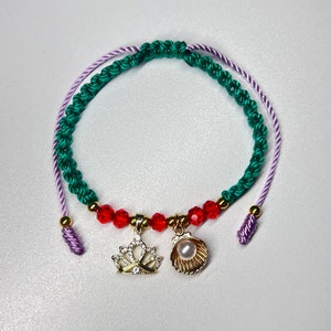 May include: A green and purple braided bracelet with red beads and gold charms. The charms are a crown and a seashell.
