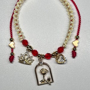Könnte beinhalten: Ein geflochtenes Armband in Rot, Weiß und Blau mit goldenen Anhängern, darunter eine Krone, ein Herz und eine Rose in einem Käfig. Das Armband ist mit roten Perlen verziert.