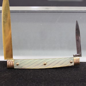 May include: A small pocket knife with a white mother-of-pearl handle and gold accents. The blade is closed and the knife is displayed in a glass case.