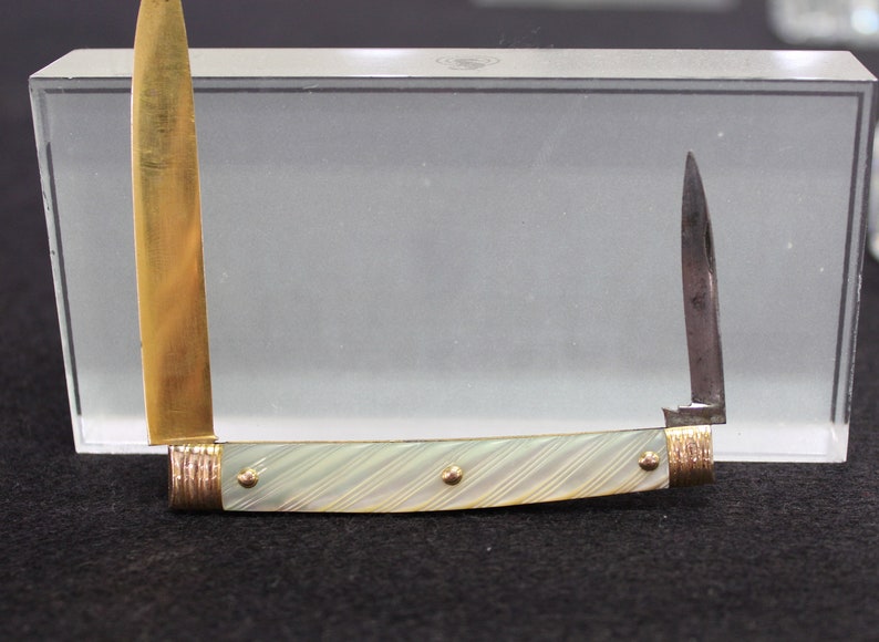 May include: A small pocket knife with a white mother-of-pearl handle and gold accents. The blade is closed and the knife is displayed in a glass case.