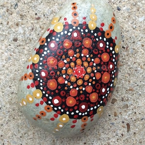 Puede incluir: Una piedra pintada con un diseño de mandala negro, naranja, rojo y blanco.