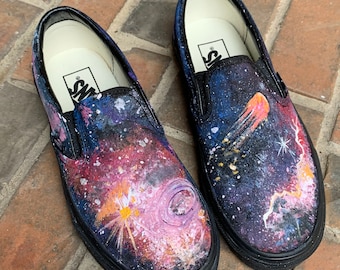 Puede incluir: Un par de zapatos Vans negros de slip-on pintados con un diseño de galaxia. Los zapatos presentan un fondo azul oscuro con estrellas blancas, una nebulosa rosa y un cometa amarillo.
