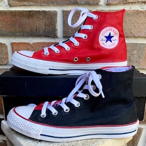 Puede incluir: Un par de zapatillas Converse de caña alta. Una zapatilla es roja con cordones blancos y el logotipo de Converse All Star. La otra zapatilla es negra con cordones blancos y una franja roja alrededor de la parte superior.