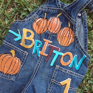 Puede incluir: Overol de mezclilla azul con un diseño pintado a mano con calabazas y el nombre "Britton" en letras de colores. El overol también tiene un número "1" pintado en el bolsillo.