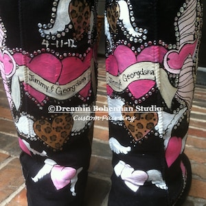Puede incluir: Un par de botas negras Ugg con diseños pintados a medida. Las botas presentan corazones rosas, alas blancas, estampado de leopardo y pedrería. El texto "Jimmy & Georgdana" y la fecha "4-11-12" están pintados en las botas.