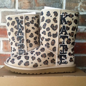 Puede incluir: Un par de botas de color marrón y negro con estampado de leopardo con el nombre "KATIE" escrito en el lateral de cada bota.