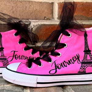 May include: A pair of bright pink high-top trainers featuring a black graphic of the Eiffel Tower and the word "Journey" in black script. The shoes have white rubber toe caps, white soles with black stripes, and black tulle ribbon shoelaces.