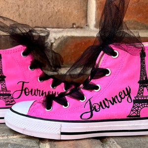 May include: Pink high-top canvas sneakers with black laces and a black tulle bow. The shoes feature a black silhouette of the Eiffel Tower and the word "Journey" written in cursive.